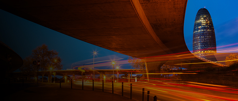 
				Modern skyscraper illuminated at night in Barcelona, seen from under a bridge.
			